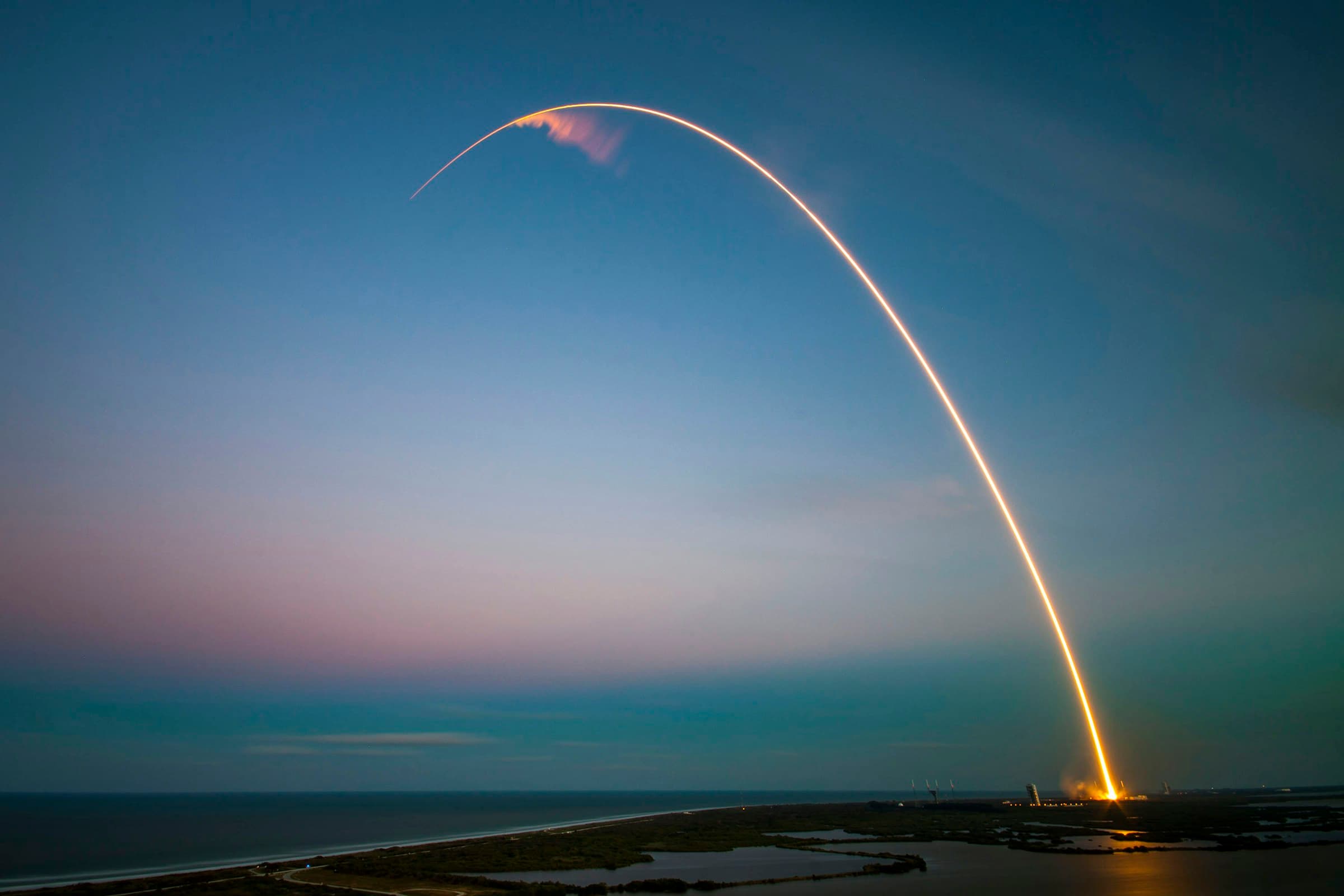 A rocket's launch trail arcing across a dusk sky over the coast